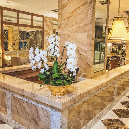 Windsor Marapendi Hotel Рио де Жанейро Екстериор снимка The photo depicts a luxurious hotel lobby featuring a polished marble reception desk. There are elegant white orchids arranged in a gold pot on the counter, adding a touch of sophistication. The surrounding interior showcases marble walls, stylish li
