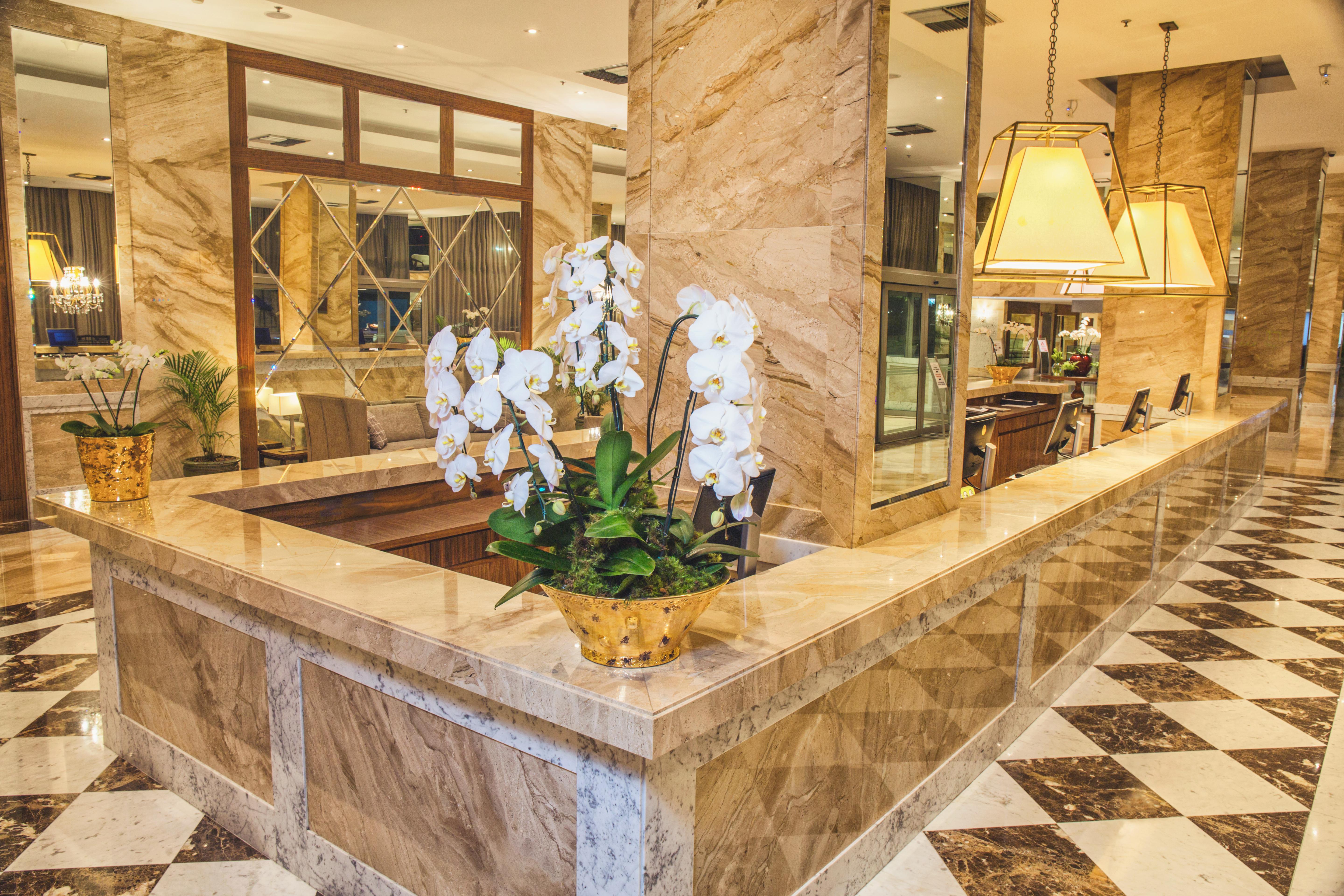 Windsor Marapendi Hotel Рио де Жанейро Екстериор снимка The photo depicts a luxurious hotel lobby featuring a polished marble reception desk. There are elegant white orchids arranged in a gold pot on the counter, adding a touch of sophistication. The surrounding interior showcases marble walls, stylish li