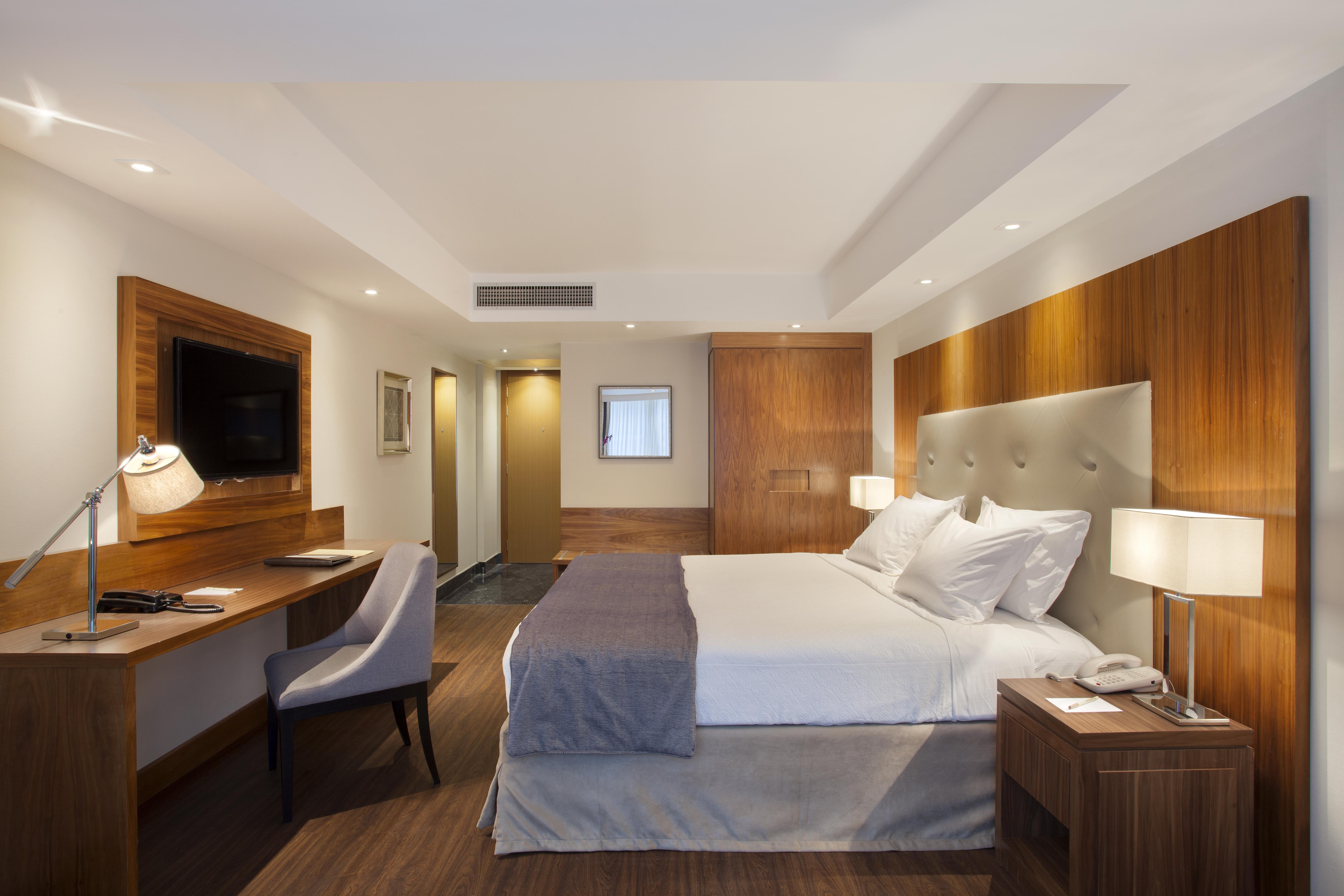 Windsor Marapendi Hotel Рио де Жанейро Екстериор снимка The photo shows a modern hotel room featuring a large bed with white and gray bedding. The headboard is upholstered in light fabric. There are two bedside lamps on either side of the bed, providing warm lighting. A television is mounted on the wall o